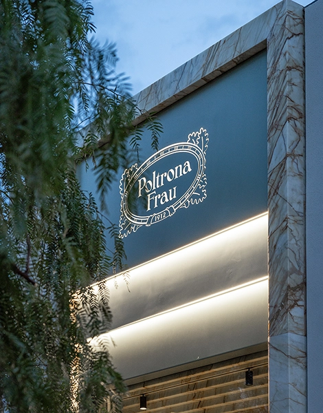 Insegna luminosa Poltrona Frau su facciata moderna in marmo, vista dal basso al crepuscolo, con rami di alberi in primo piano.
